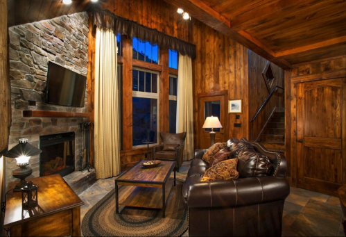 Cozy living room with wooden walls, stone fireplace, large windows, and rustic furniture. Warm, inviting atmosphere.