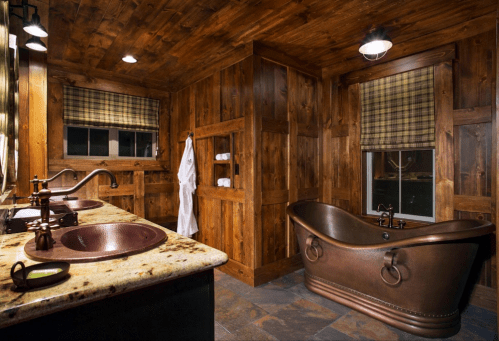 Cozy rustic bathroom with wooden walls, a copper bathtub, and dual sinks, featuring warm lighting and a plush robe.