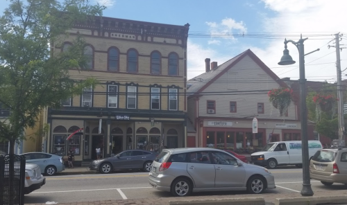 Historic buildings line a street, featuring shops and restaurants, with cars parked along the road under a blue sky.
