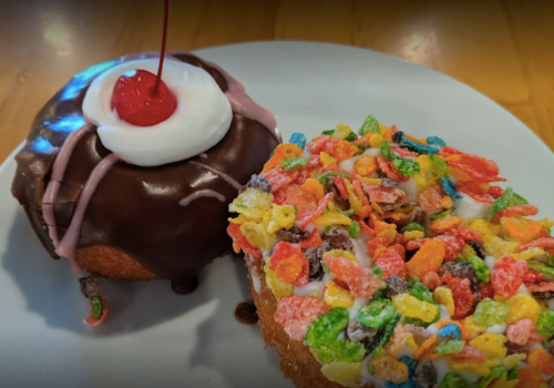 A plate with two colorful donuts: one topped with chocolate icing and a cherry, the other with fruity cereal.