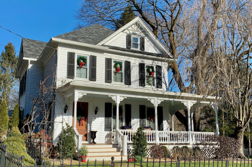 A charming white house with black shutters, decorated with holiday wreaths, surrounded by trees and a picket fence.