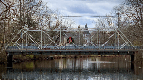 A metal bridge spans a calm river, adorned with a wreath, with a church steeple visible in the background.