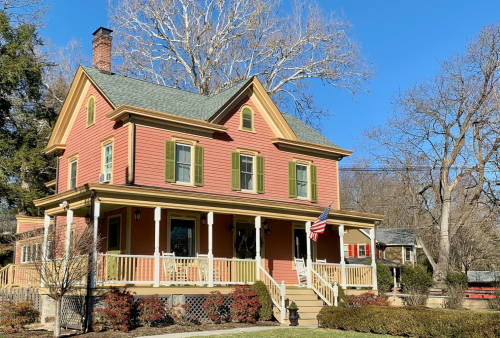 A charming pink Victorian house with green shutters, a porch, and an American flag, surrounded by trees and blue sky.