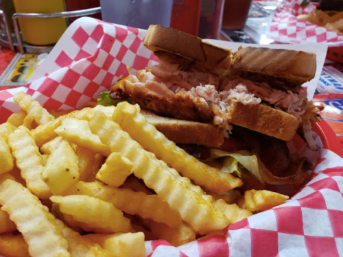 A sandwich with layers of meat and lettuce, served with a side of crinkle-cut fries in a red-checkered basket.