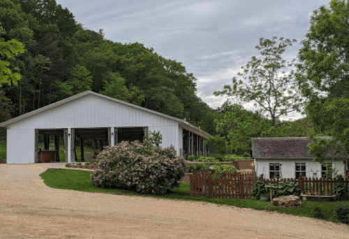 A white barn and a small house surrounded by greenery and flowers, with a gravel path leading to them.