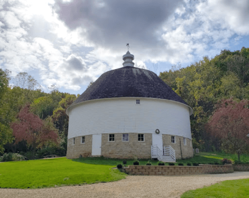 A round white building with a dark roof, surrounded by greenery and trees under a partly cloudy sky.