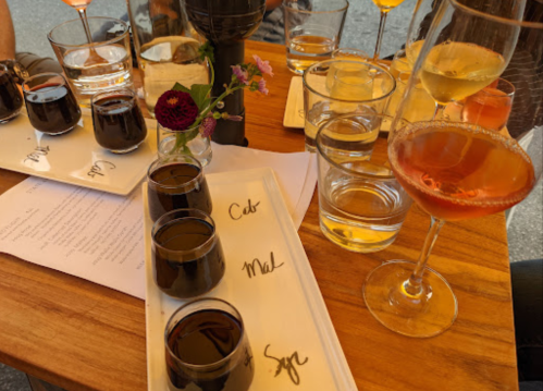 A wooden table with various glasses of drinks, including dark and light beverages, and a small flower centerpiece.