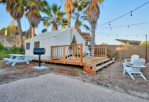 A glamping tent on a wooden deck surrounded by palm trees, with a picnic table and seating area nearby.