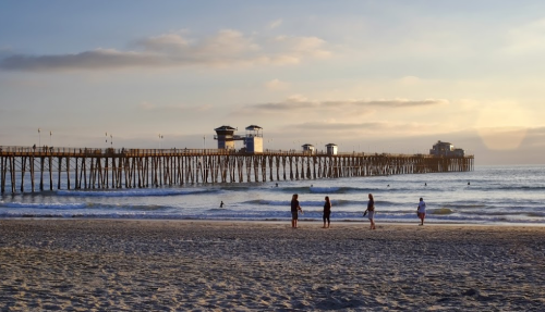 A sandy beach at sunset with a wooden pier in the background and people walking along the shore.