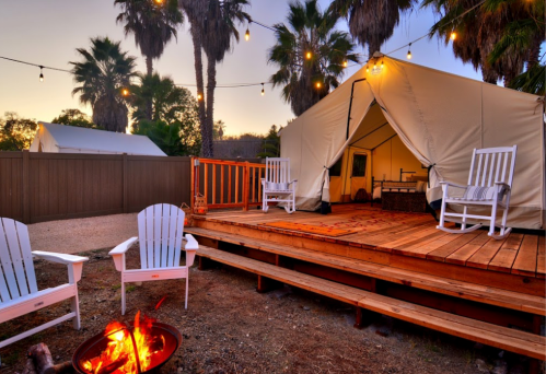 A cozy glamping tent on a wooden deck, surrounded by palm trees and string lights, with a fire pit in front.