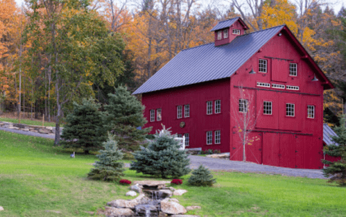 A red barn surrounded by green grass, evergreen trees, and autumn foliage, with a small stream in the foreground.