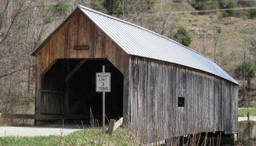 A rustic wooden covered bridge with a metal roof, featuring a weight limit sign, surrounded by trees and hills.