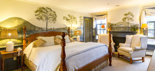 Cozy bedroom with a wooden bed, armchairs, and scenic wall murals of hills and trees. Warm lighting enhances the ambiance.