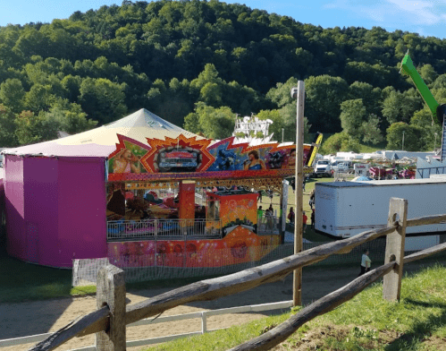 A colorful carnival ride with a tent, surrounded by trees and a fairground in the background. A wooden fence in the foreground.