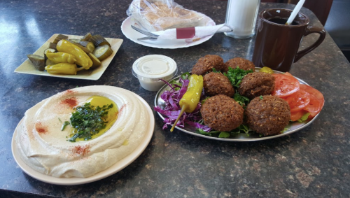 A plate of falafel with salad, hummus drizzled with olive oil, pickles, and a cup of coffee on a table.