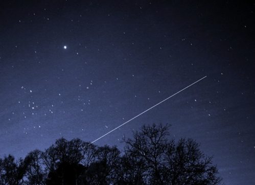 A night sky filled with stars, featuring a bright object and a streak of light, possibly a satellite, above silhouetted trees.