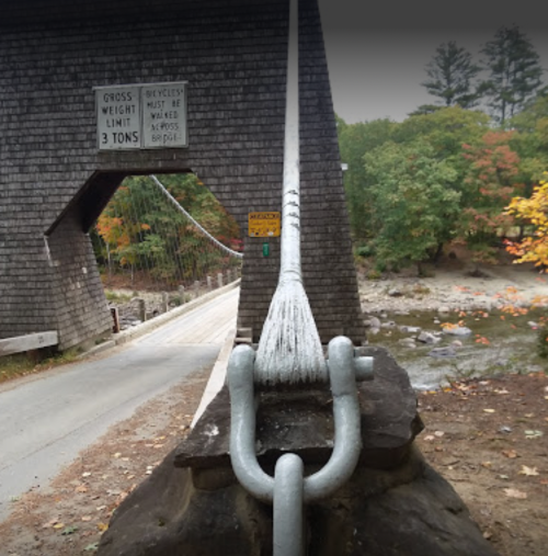 A cable supports a wooden bridge with a sign indicating weight limit and bicycle restrictions, surrounded by autumn foliage.