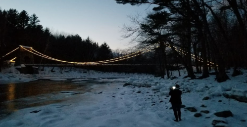 A person stands by a snowy riverbank, illuminated by string lights on a bridge in the background at dusk.