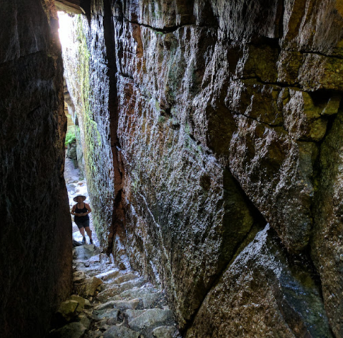 A narrow stone passageway with steep steps, leading to a person standing at the bottom, surrounded by mossy rock walls.