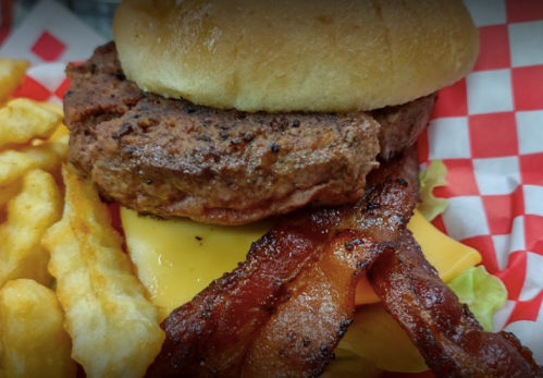 A close-up of a burger with a beef patty, cheese, bacon, and crinkle-cut fries on a red and white checkered tray.