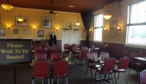A cozy restaurant interior with empty tables and chairs, featuring a "Please Wait To Be Seated" sign.