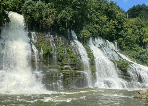 A cascading waterfall surrounded by lush greenery under a clear blue sky. Water flows powerfully over rocky cliffs.