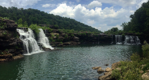 A scenic view of cascading waterfalls surrounded by lush greenery and a calm lake under a partly cloudy sky.