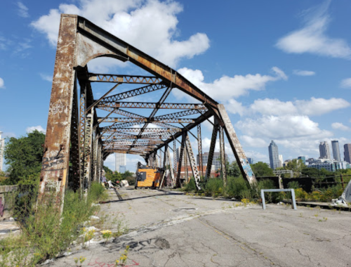 Rusty, abandoned bridge with overgrown vegetation and a city skyline in the background under a blue sky with clouds.