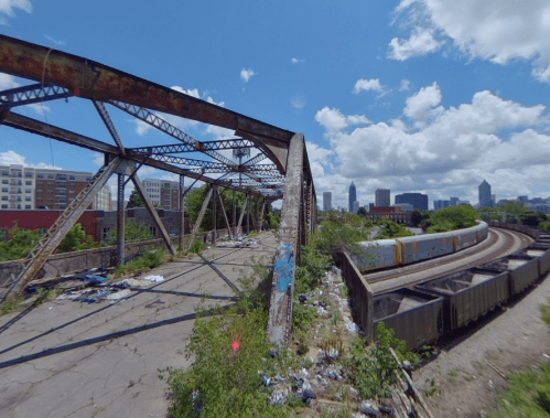 Abandoned bridge with rusted metal structure, overgrown vegetation, and a view of a city skyline under a blue sky.
