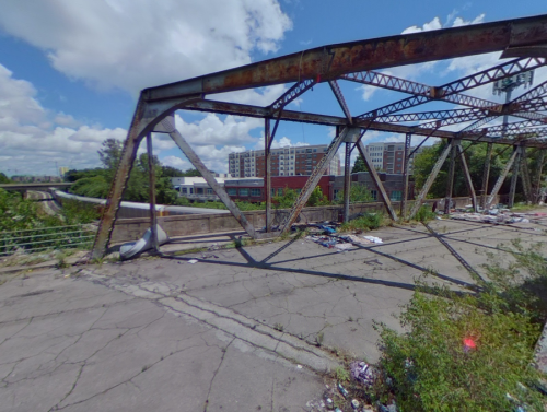 Abandoned bridge structure with rusted metal beams, overgrown vegetation, and scattered debris, set against a city backdrop.