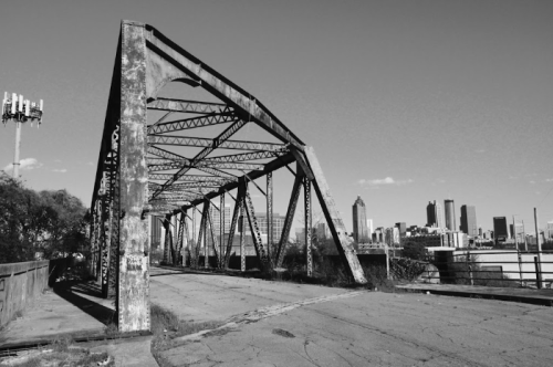 Black and white image of a weathered bridge with a city skyline in the background.