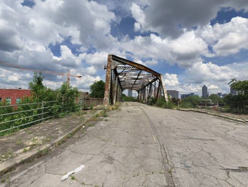 An old, rusty bridge spans a cracked road, surrounded by overgrown vegetation and a cloudy sky.