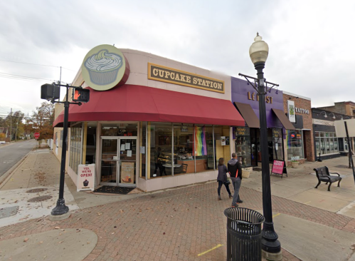 A storefront of Cupcake Station with a red awning, featuring a cupcake sign and nearby shops on a street corner.