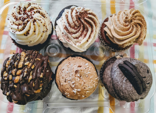 A variety of six cupcakes with different frosting styles and toppings, displayed in a clear container.