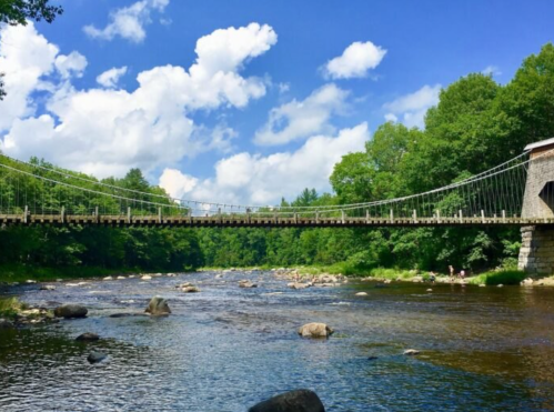 A scenic view of a suspension bridge over a calm river, surrounded by lush green trees and a blue sky with clouds.