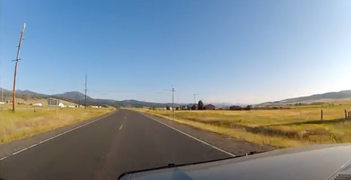 A scenic view of a rural road with fields and mountains under a clear blue sky.