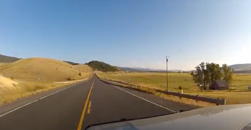 A scenic view of a long, empty road stretching through golden fields and rolling hills under a clear blue sky.
