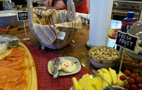 A buffet table with bagels, smoked salmon, fresh fruit, and a basket, set in a kitchen.