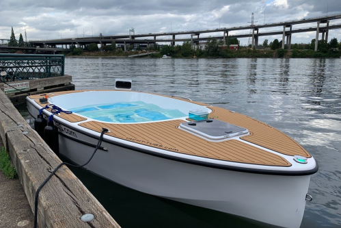 A sleek white boat with a wooden deck is docked by a river, with a bridge visible in the background under cloudy skies.