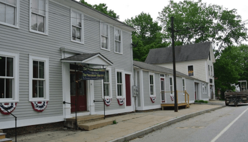 A row of historic buildings with decorative bunting, set along a quiet street lined with trees.