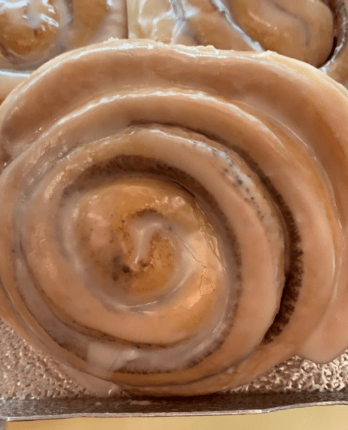 Close-up of a glazed cinnamon roll with a golden-brown swirl and creamy icing on top.