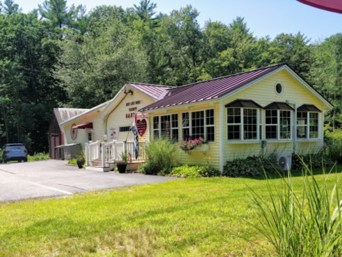 A charming yellow house with a red roof, surrounded by greenery and a gravel driveway.