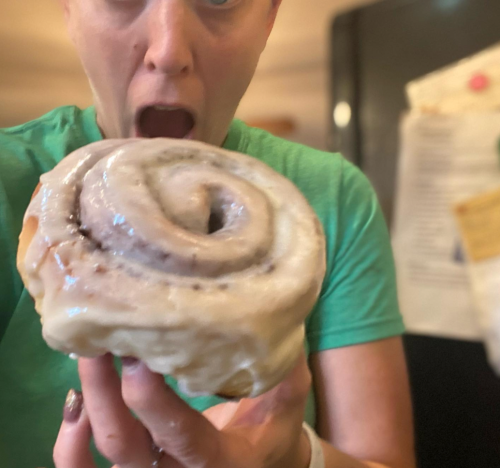 A person in a green shirt excitedly holds a large cinnamon roll, mouth open in surprise.