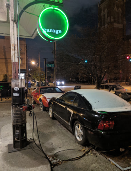 A neon green "Garage" sign illuminates two parked cars, one covered in frost, at a gas station at night.