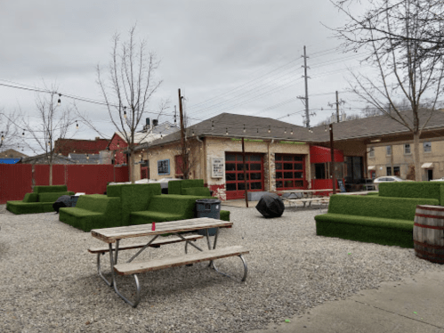 Outdoor space with green turf seating, picnic tables, and a building in the background under a cloudy sky.