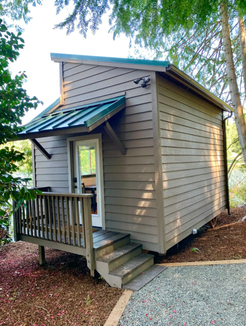 A small, modern cabin with a green roof, wooden steps, and surrounded by trees and gravel pathways.