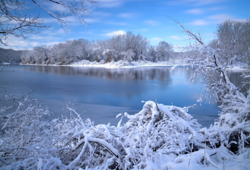 A serene winter landscape featuring a snow-covered riverbank and trees, reflecting a clear blue sky.
