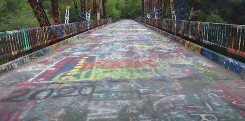 A colorful, graffiti-covered bridge with vibrant designs and messages on the pavement, surrounded by greenery.