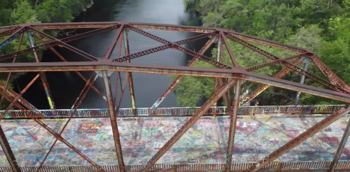 Aerial view of a rusty, graffiti-covered bridge over a calm river surrounded by lush greenery.
