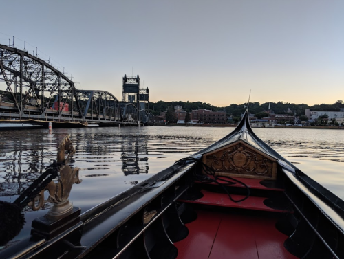 View from the front of a gondola on calm water, with a city skyline and bridge in the background at sunset.